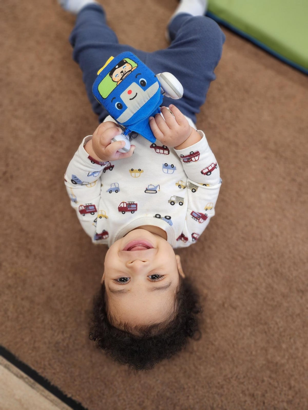 Smiling baby lying on a carpet and holding a blue toy vehicle, looking up at the camera.