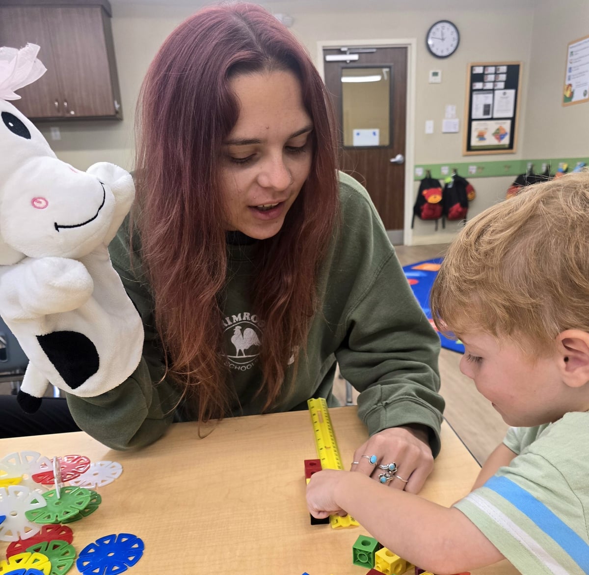 Toddler measuring blocks with a teacher using a tape measure during a hands-on learning activity at a daycare in Keller, Texas, with a cow puppet used for playful engagement.