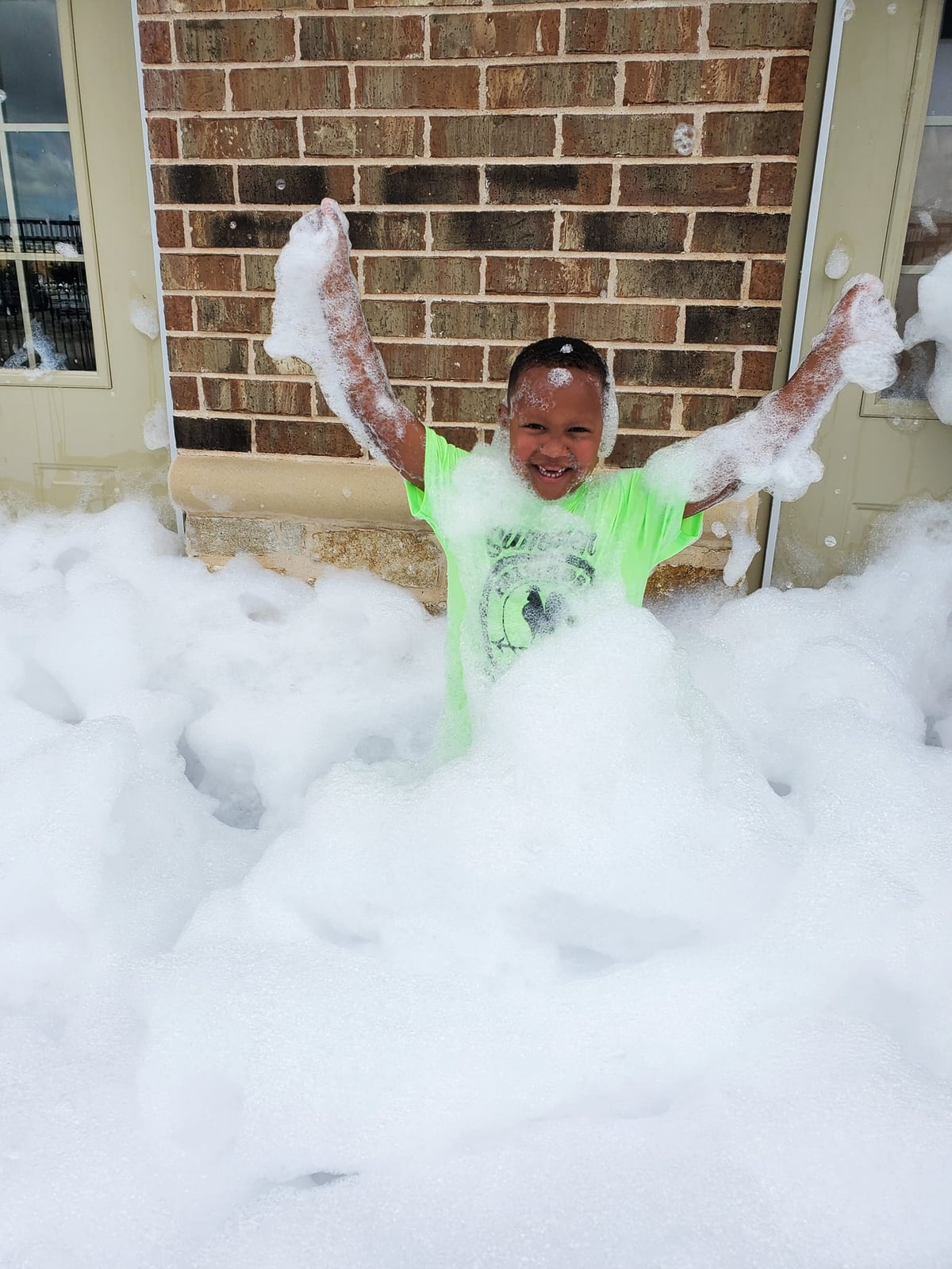Primrose School summer camper playing in thick white foam outdoors during a Foam Day activity, celebrating summer fun and sensory play with big smiles and laughter at the best summer camp in Haslet. 