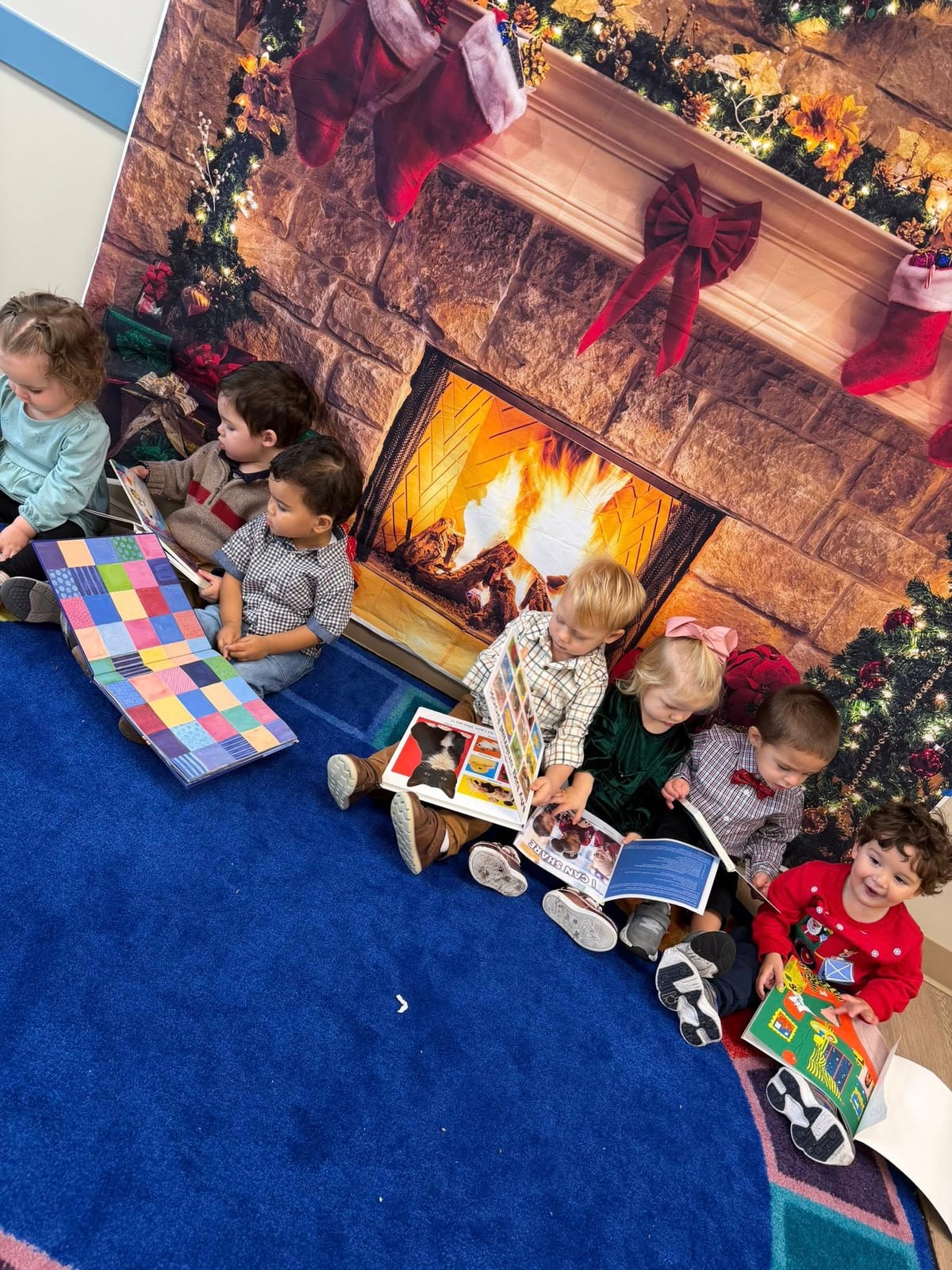 children sitting in front of a fireplace backdrop on a blue carpet reading books 