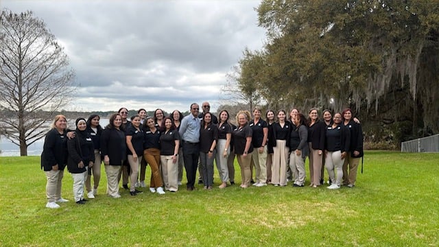 Group of teachers in khaki pants and black polos on green grass in front of a tree and lake 
