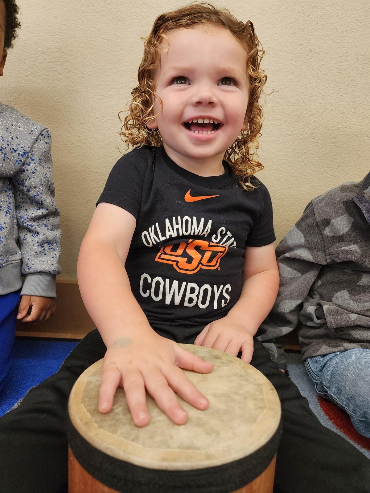 Toddler boy smiling while sitting on the carpet, beating the drum to the beat in a song during music time