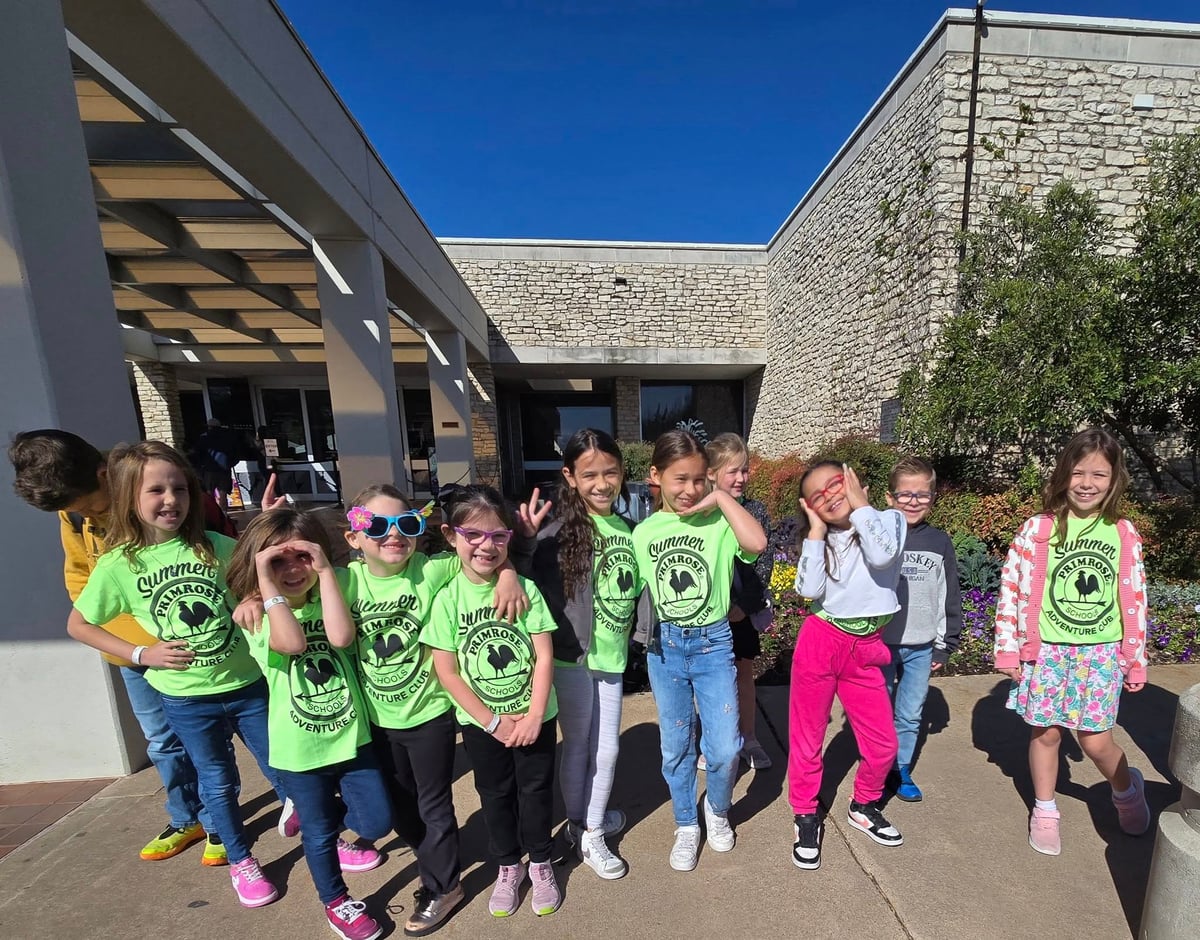 Group of preschool children wearing matching green t-shirts standing outside by a school building on a sunny day.