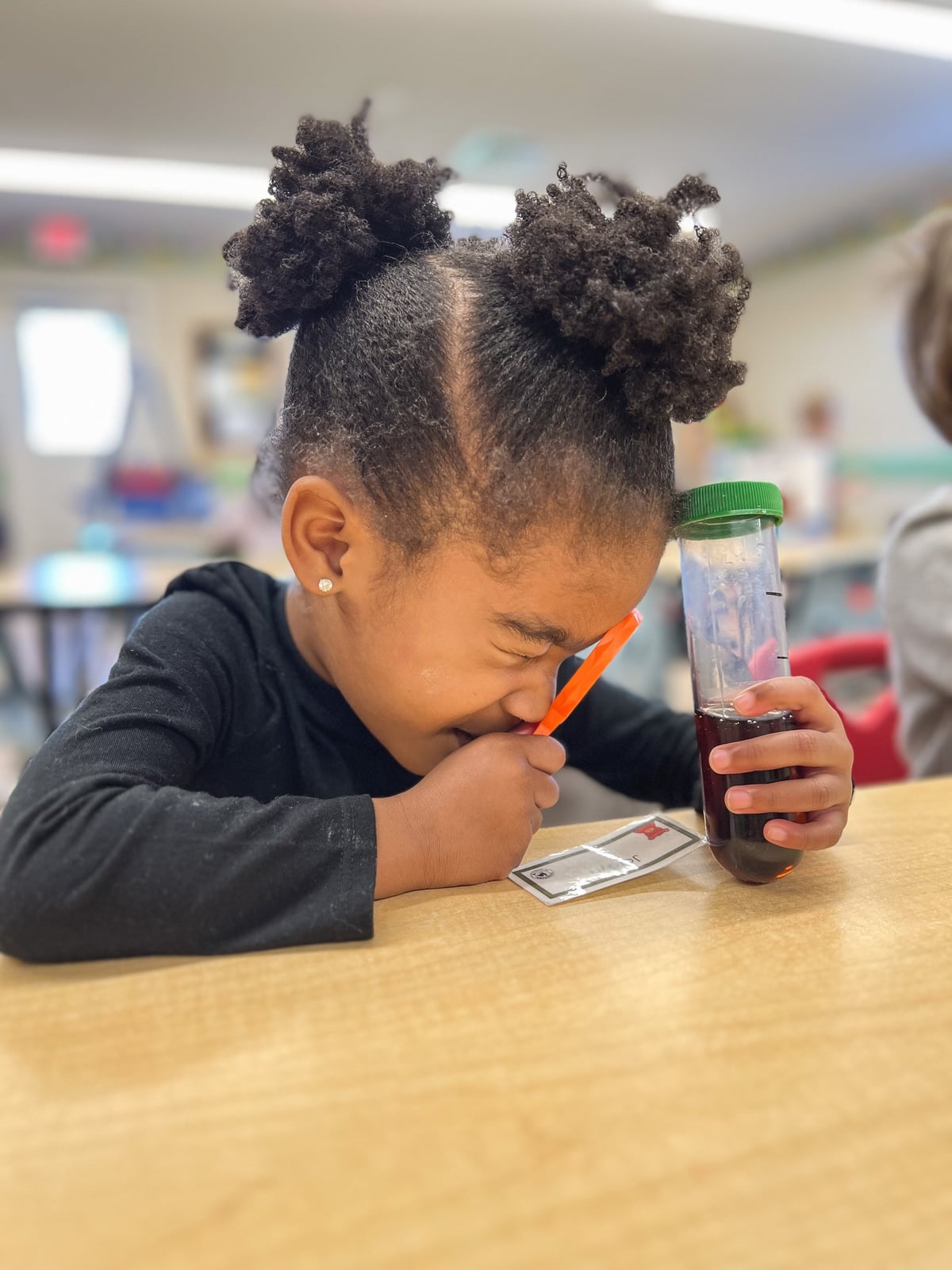 Young preschool girl exploring a sensory science activity at Primrose School of NW Fort Worth, using a magnifying glass to observe an object in a discovery tube. Hands-on learning, STEM education, and joyful curiosity in an early childhood classroom