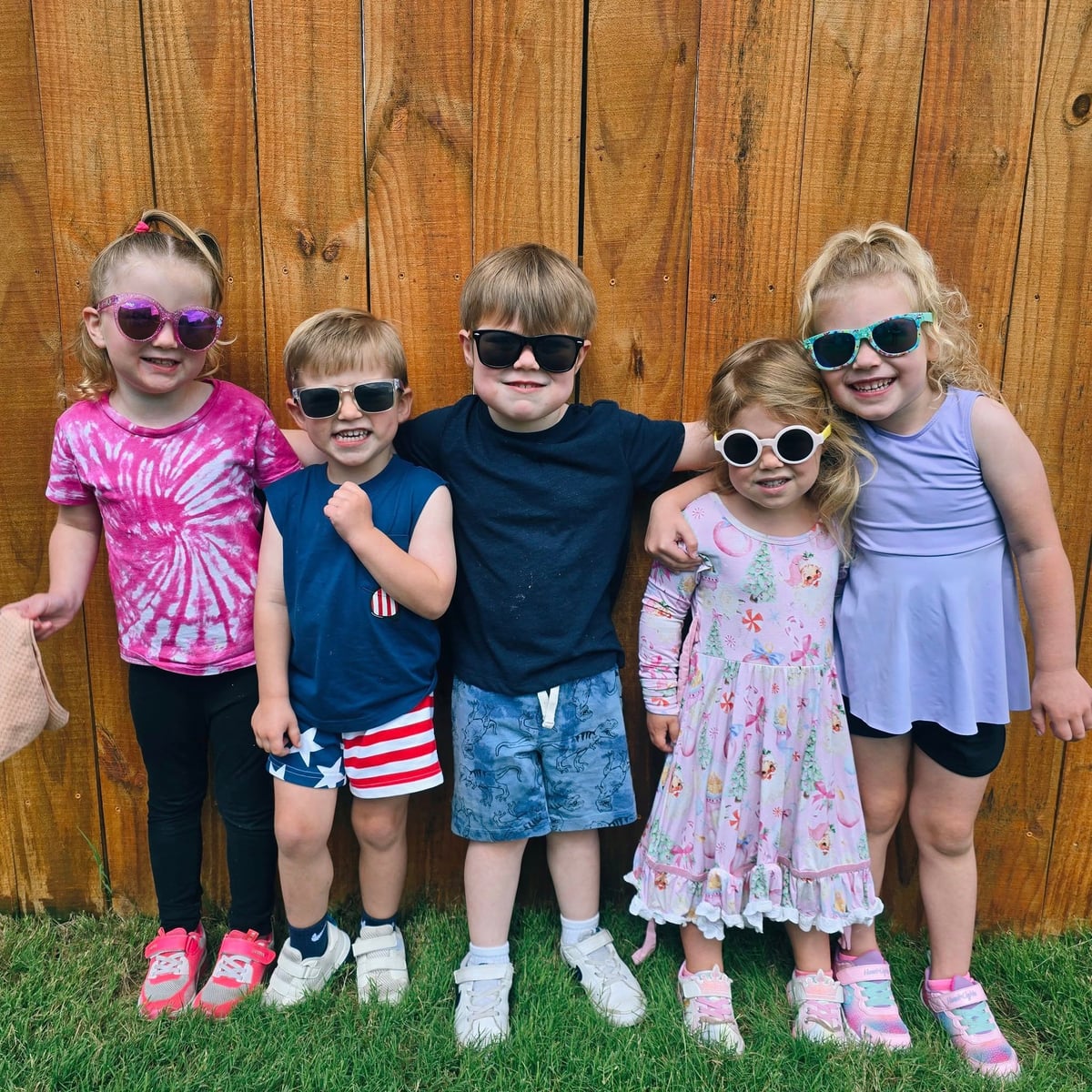 Group of preschool children wearing sunglasses and summer outfits, smiling and standing in front of a wooden fence at a childcare center in Keller, Texas.