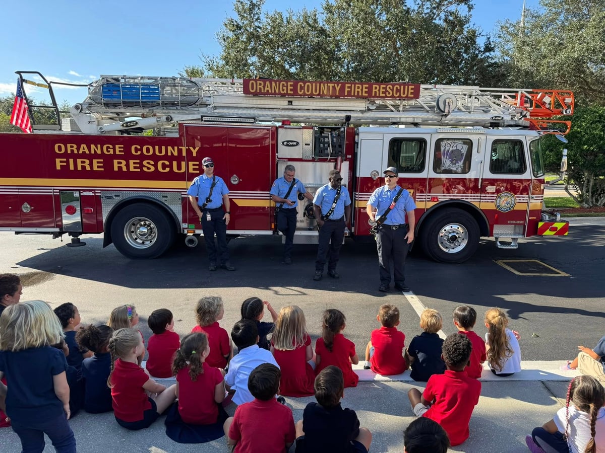children in red. white or blue shirts sitting on the curb looking at a firetruck and 4 firemen