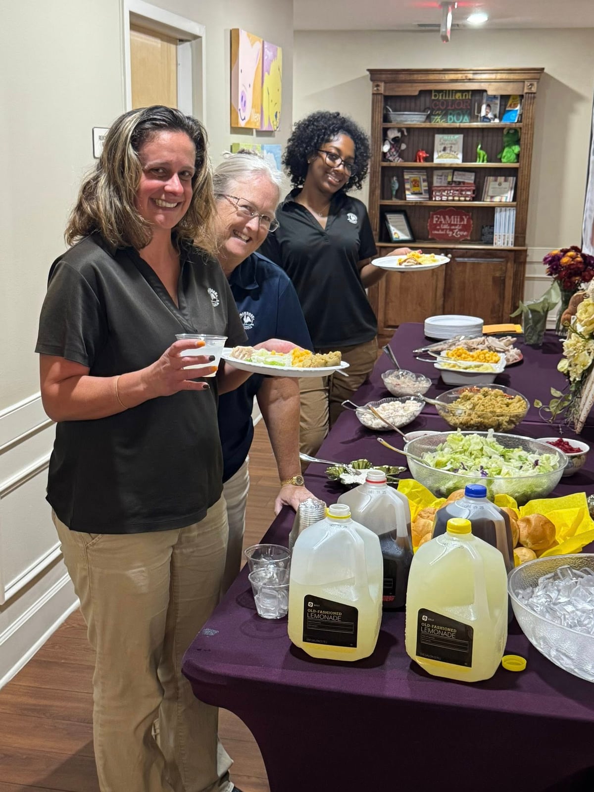three adult women in black polos and khaki pants holding plates of food standing in front of a table of food