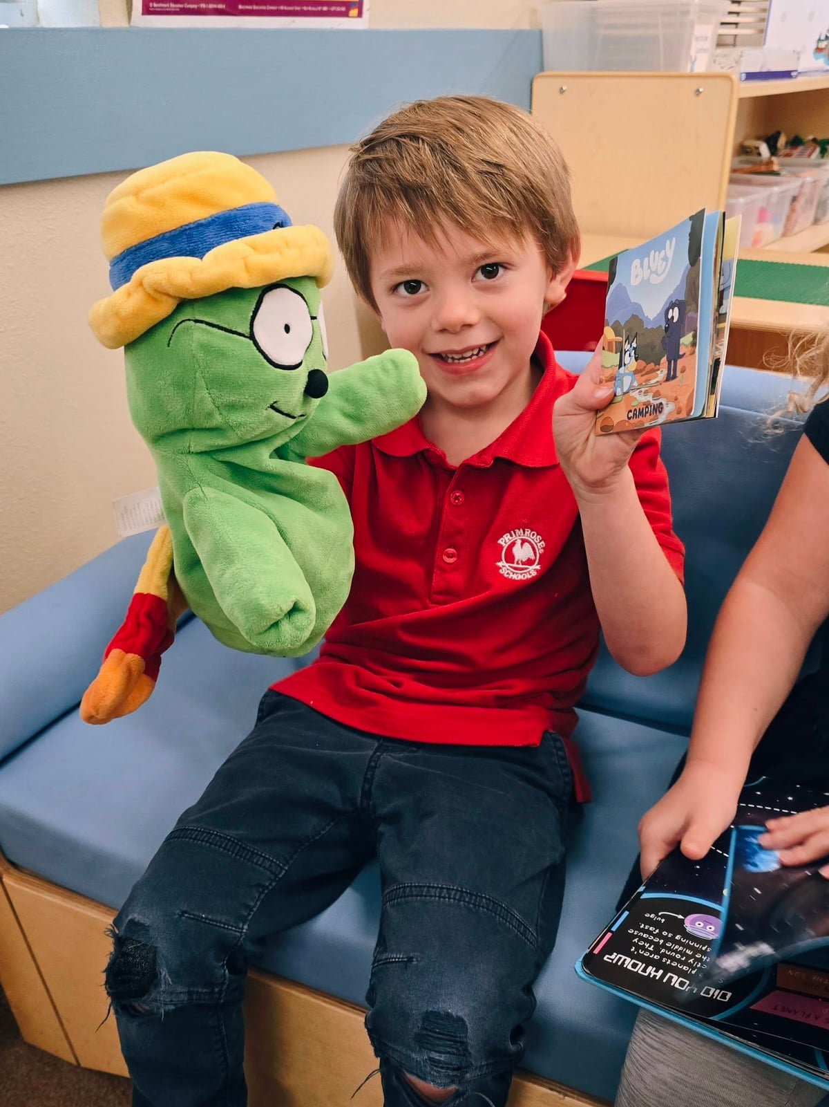 Smiling Pre-K boy holding up a book and showing it to OG the Bookworm at Primrose School at Heritage.
