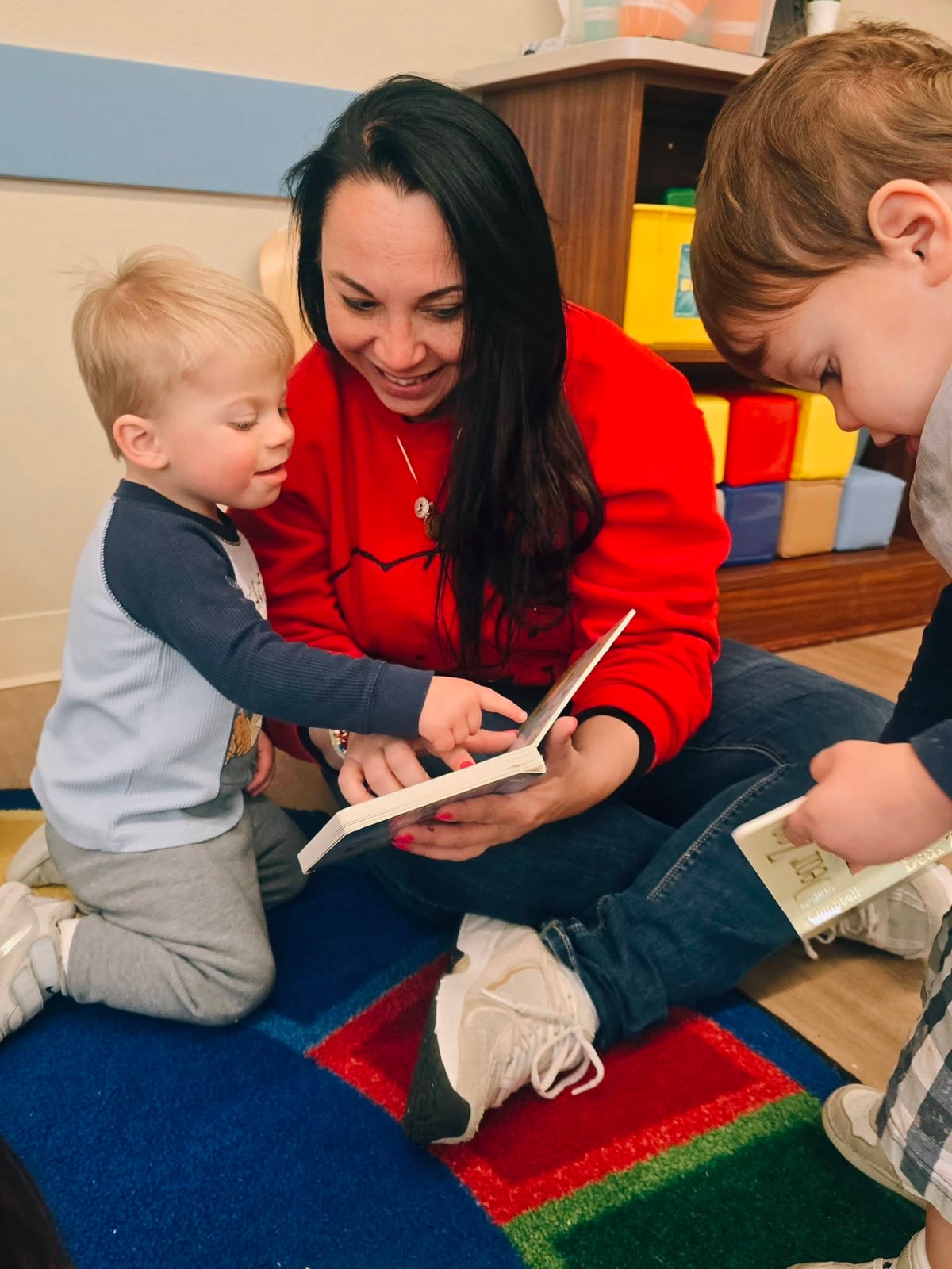 Teacher and toddler boy smiling while reading a book together in a classroom.