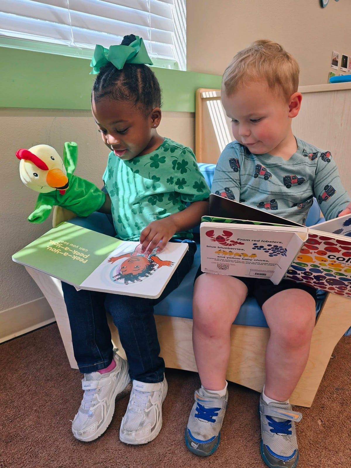 Two-year-old girl and boy sitting on a couch reading books together while the girl holds a puppet during center time at Primrose.