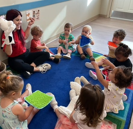 toddlers sitting on colorful rug in a circle, looking at teacher holding puppet;