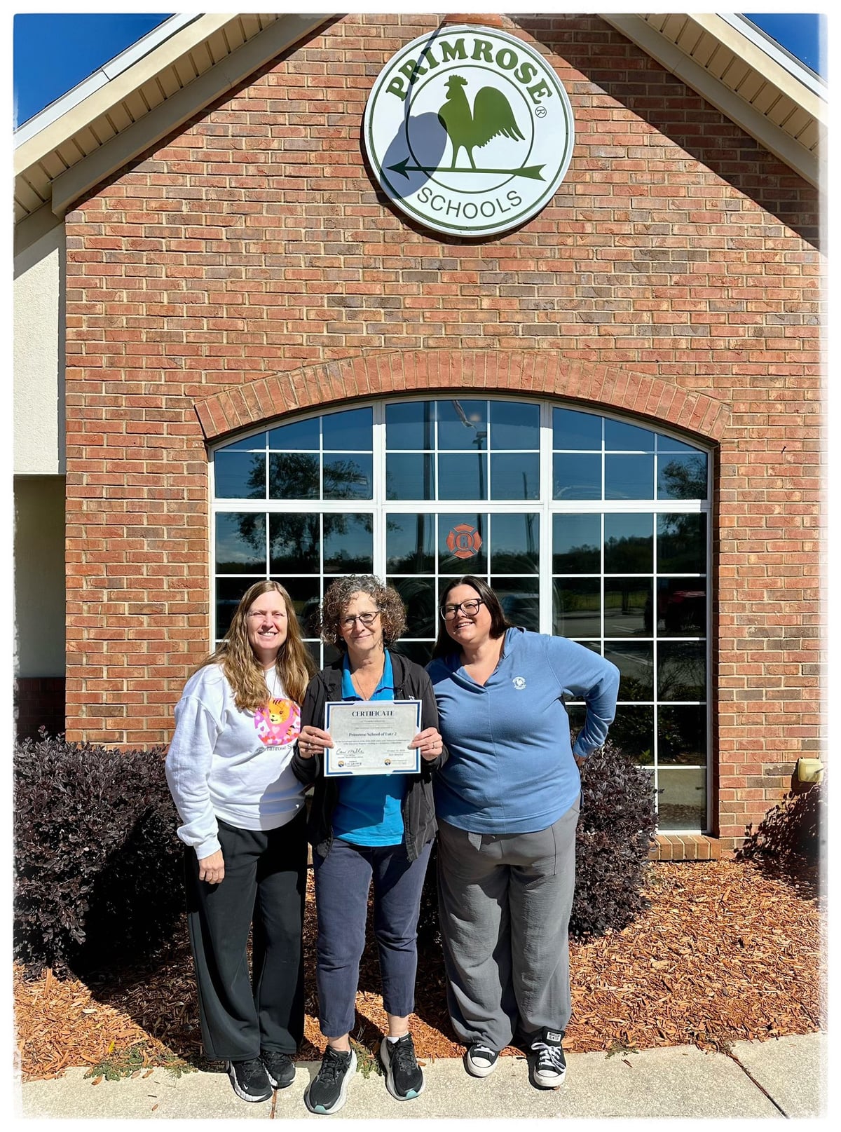 Three teachers holding a certificate in front of a building