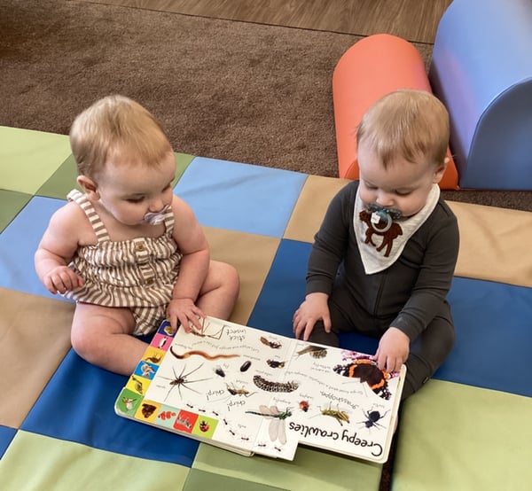 Two infants exploring board book together on mat to develop early literacy and social interaction skills.