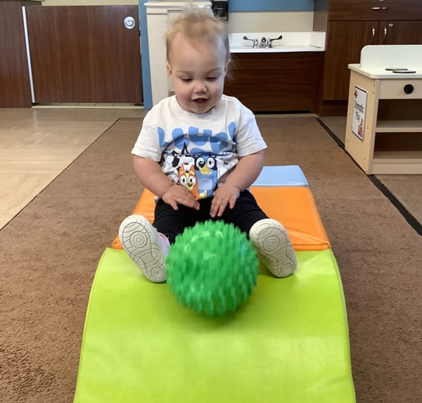 Toddler playing with a textured green ball on a soft ramp in a preschool classroom, developing gross motor skills, balance, and coordination through active play in a nurturing childcare center.