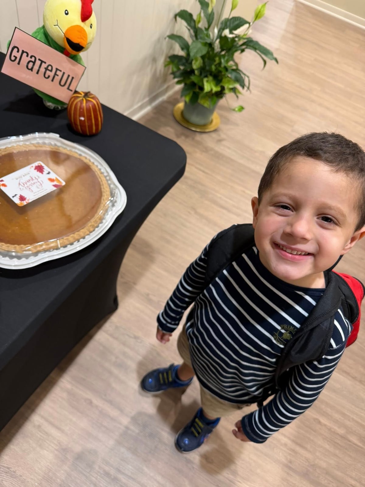 little boy in blue and white stripped shirt with a backpack on standing in front of a table with a pumpkin pie